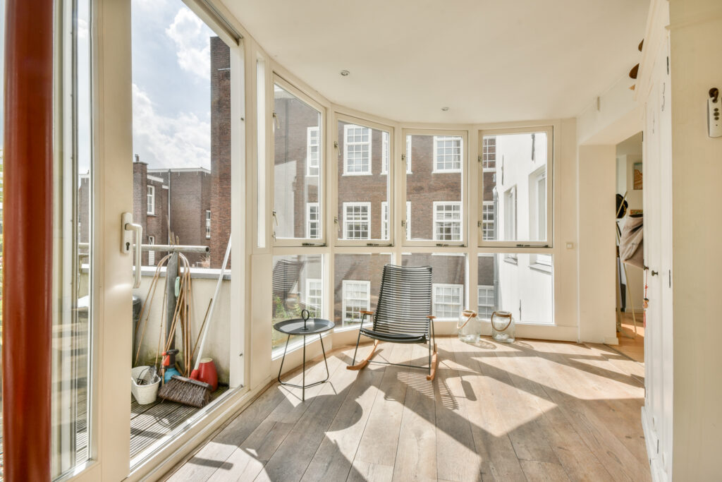 A stylish sunlit room featuring large window replacement that create a vibrant atmosphere. The minimalist decor includes a chair, small table, and wooden flooring.