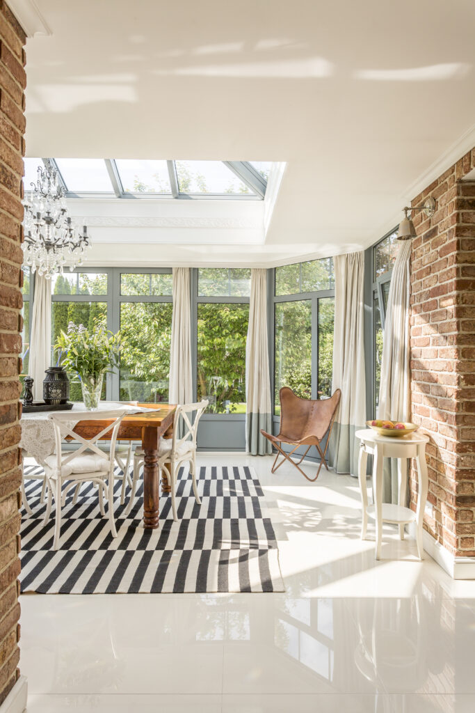 Spacious, modern dining room with picture windows around, glazed roof, stylish chandelier, brick walls, stripy carpet and table with chairs in the middle